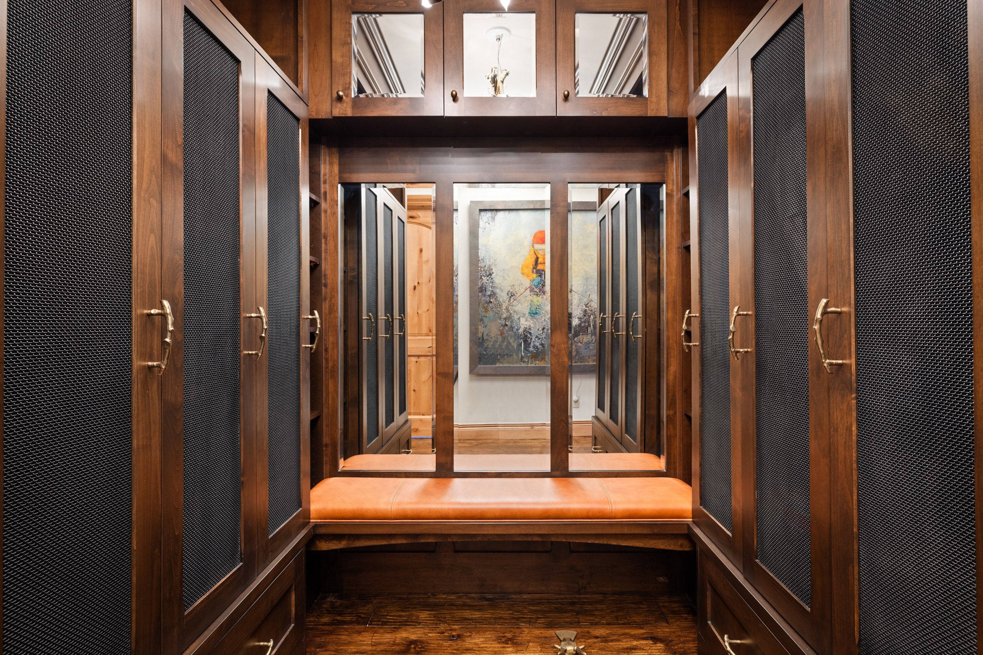 Custom-built closet system featuring rich wood cabinetry with black mesh panel inserts, brass hardware, and an integrated padded bench