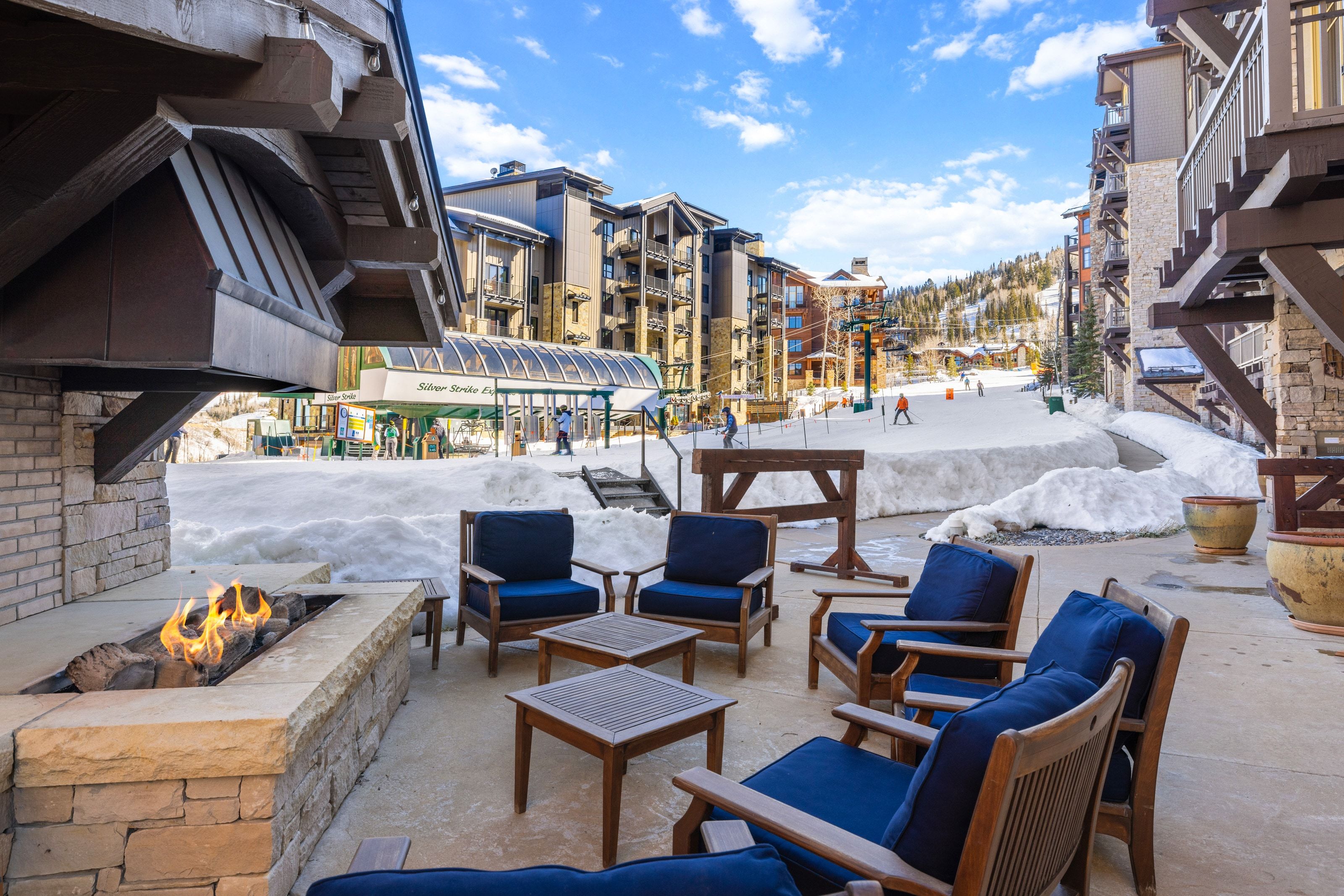 Outdoor patio featuring a stone-clad fire pit, concrete flooring, and multiple wooden chairs with blue cushions