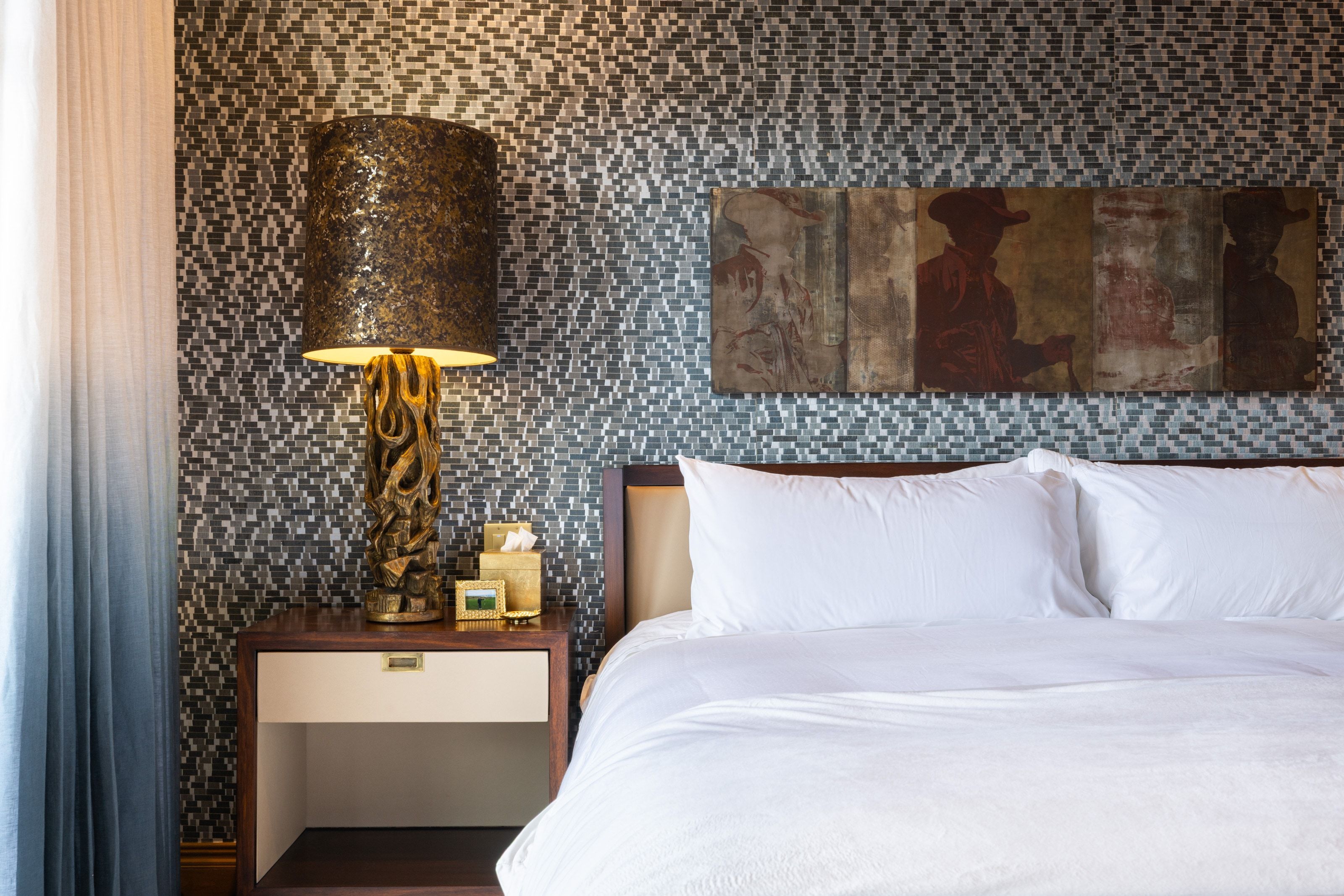 Feature wall with mosaic tile, wood-finish nightstand with single drawer, custom wall art, and a decorative table lamp