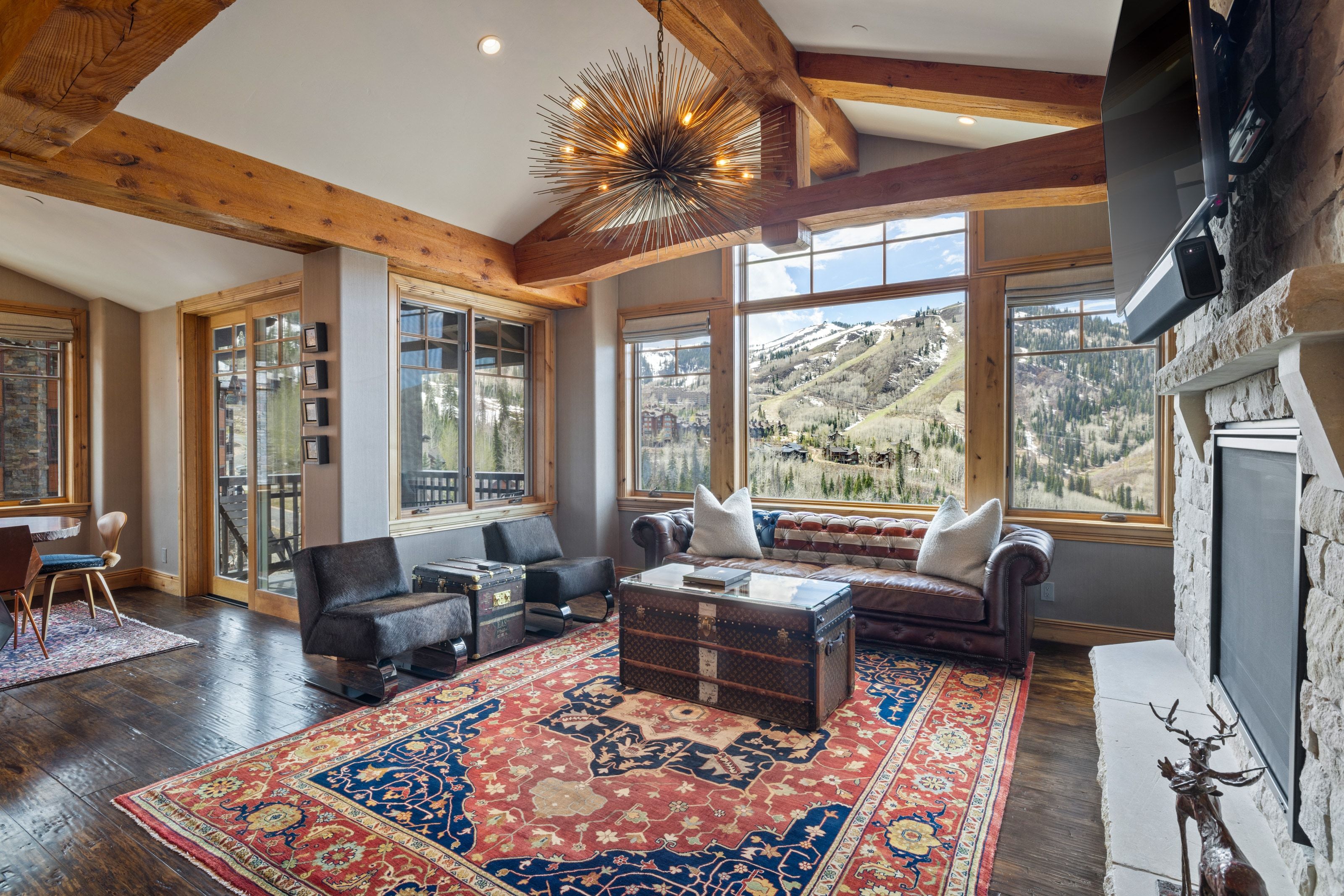 Open living area featuring expansive windows, exposed wood beams, a stone fireplace, wood-finish flooring, and a contemporary chandelier