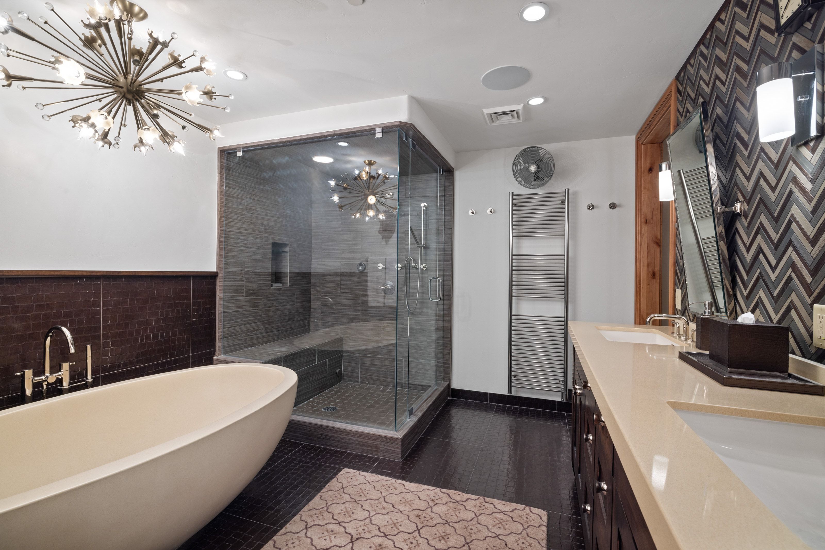 Freestanding soaking tub with floor-mounted faucet, expansive glass-enclosed shower with bench, dual vanity with undermount sinks, geometric accent wall, and contemporary chandelier