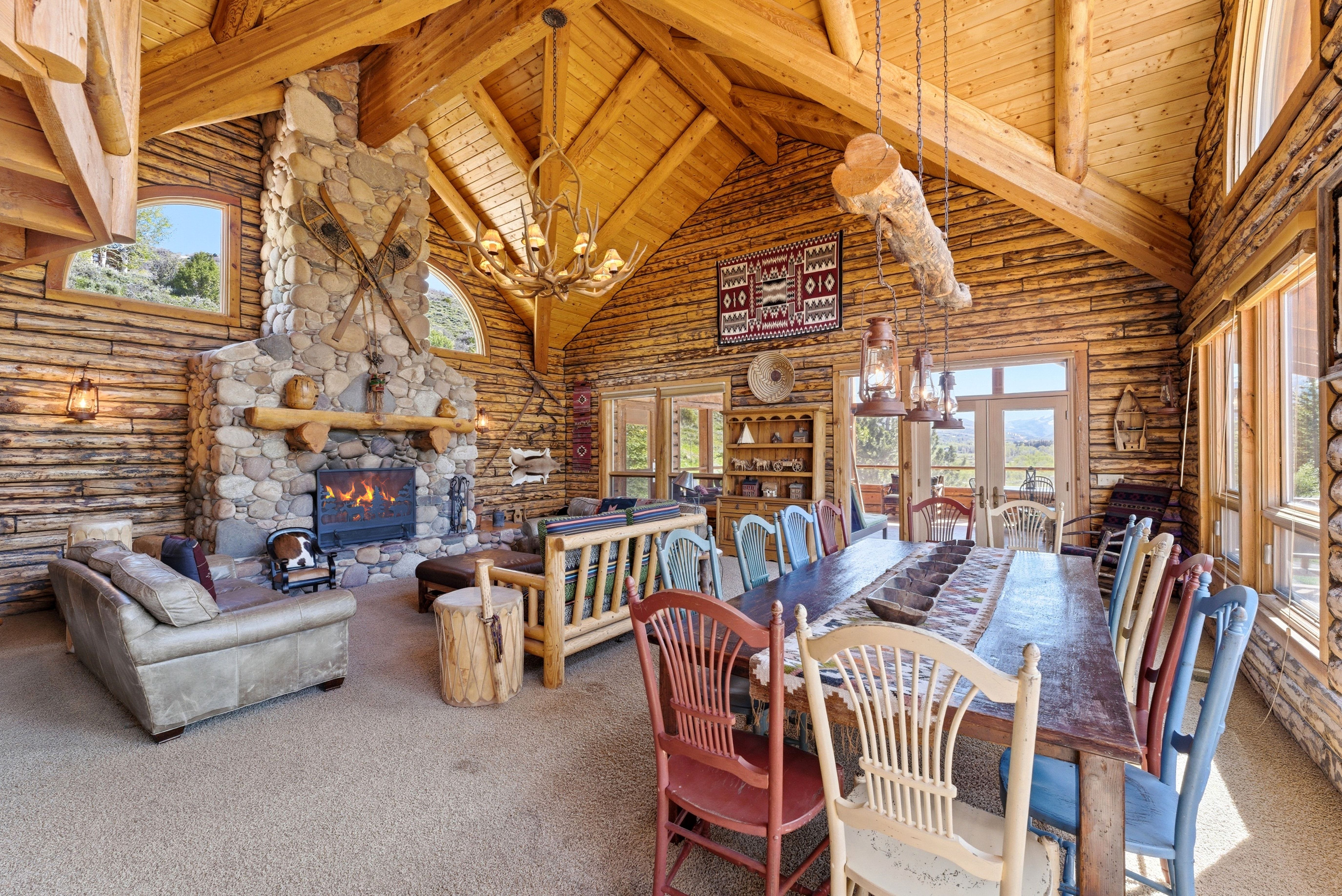 Spacious great room featuring exposed log walls, a vaulted ceiling with exposed beams, a large stone fireplace with a substantial timber mantel, a chandelier, and multiple large windows