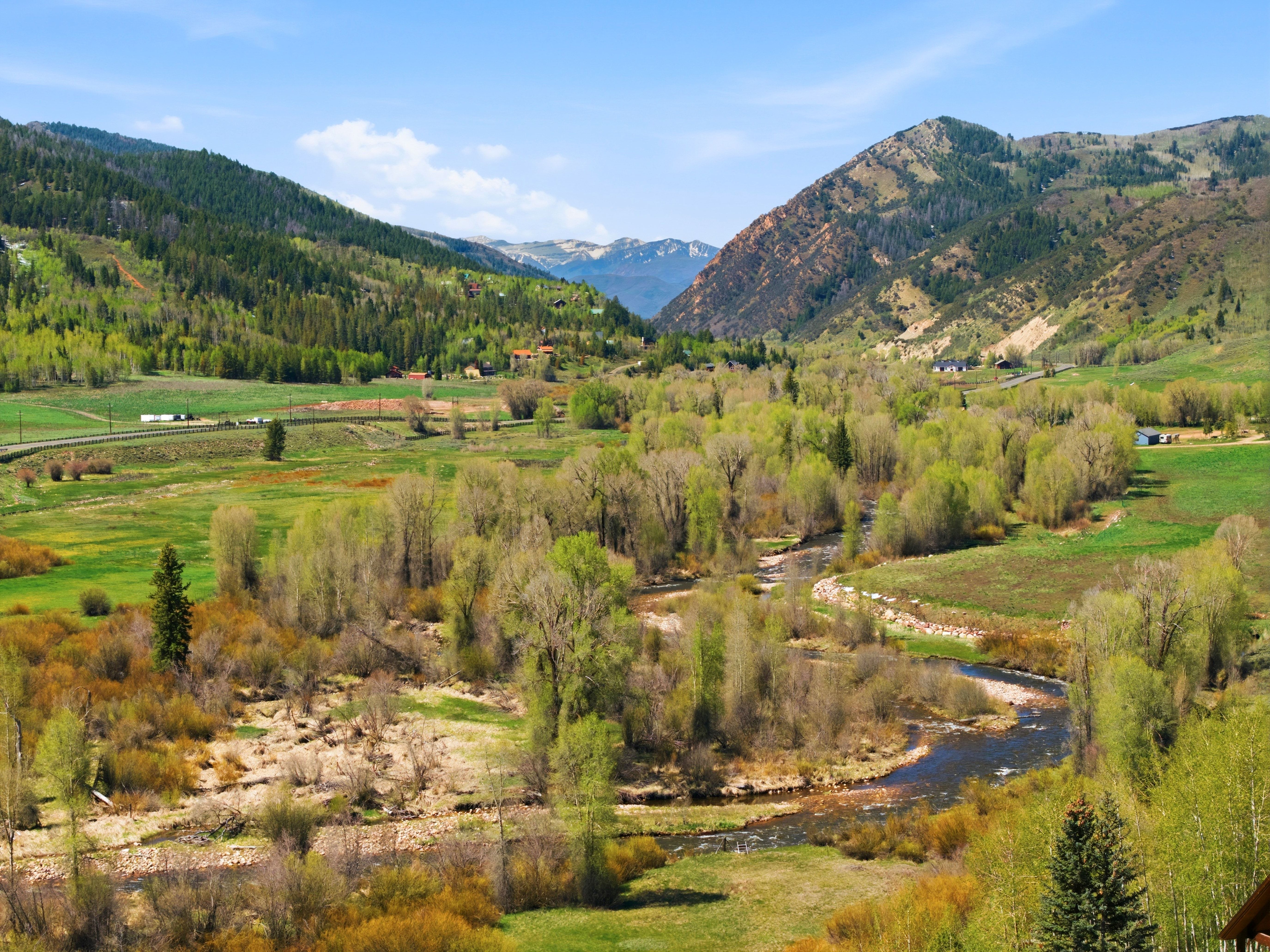 Expansive mountain valley landscape featuring a winding river, lush green fields, abundant deciduous trees, distant evergreen forests, and prominent mountain ranges