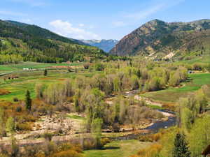 Expansive mountain valley landscape featuring a winding river, lush green fields, abundant deciduous trees, distant evergreen forests, and prominent mountain ranges