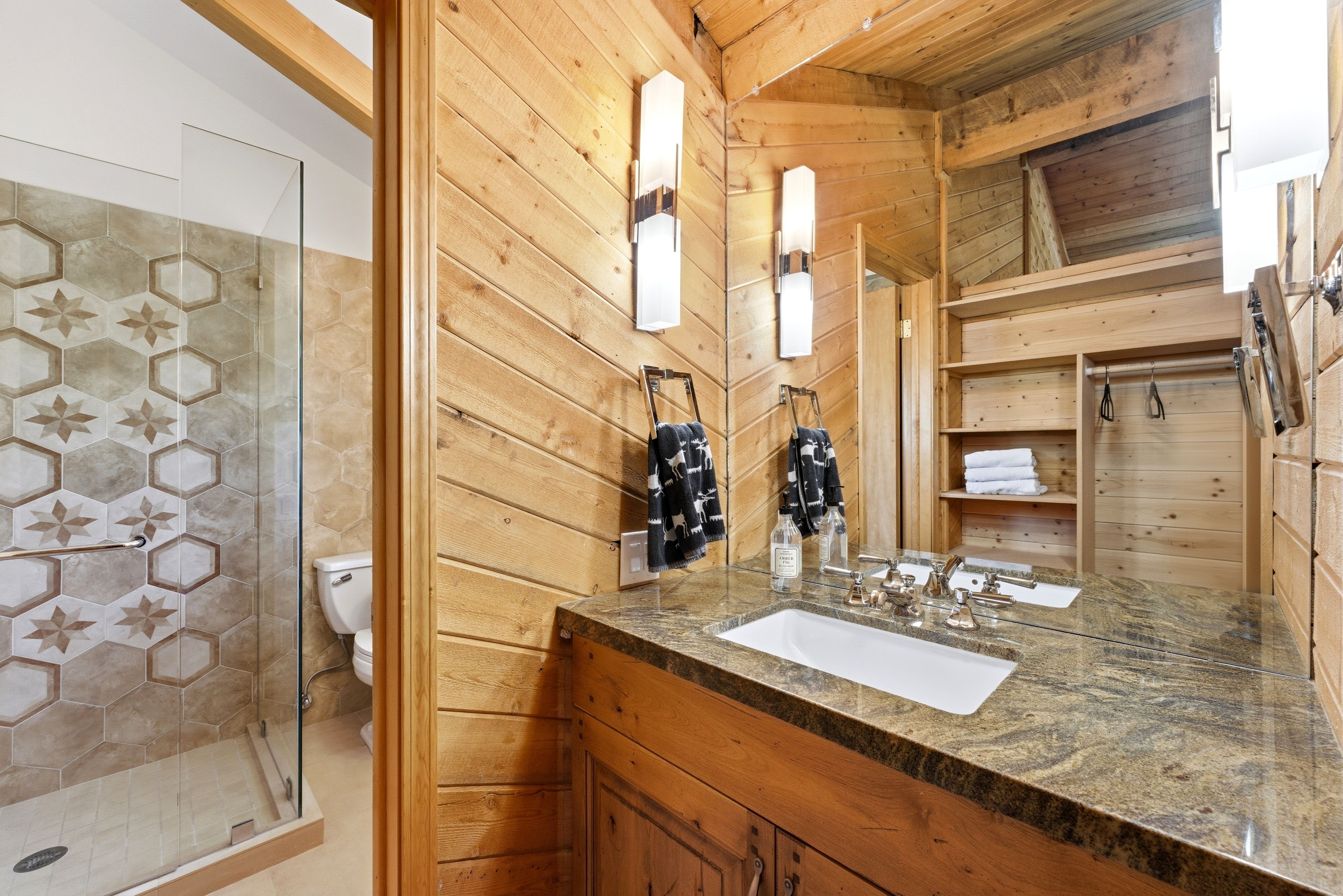 Wood-paneled bathroom featuring a granite vanity with an undermount sink, built-in shelving, glass-enclosed shower, and decorative tile accents