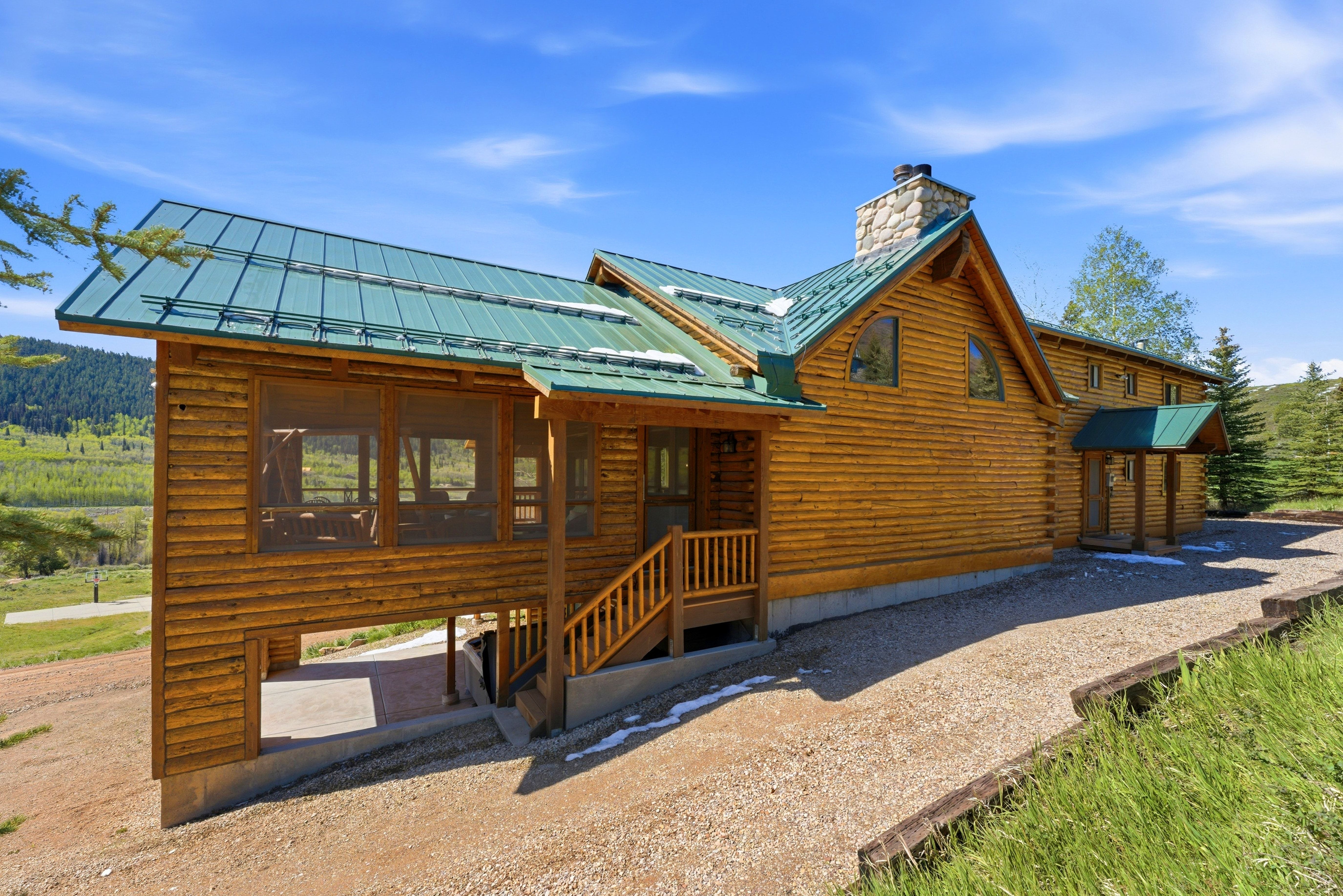 Log cabin exterior featuring a green metal roof, a screened porch, a stone chimney, a covered entryway, and a gravel driveway
