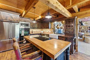 Kitchen featuring extensive wood cabinetry, stainless steel appliances, a center island with a cooktop, a farmhouse-style sink, and exposed wood ceiling beams