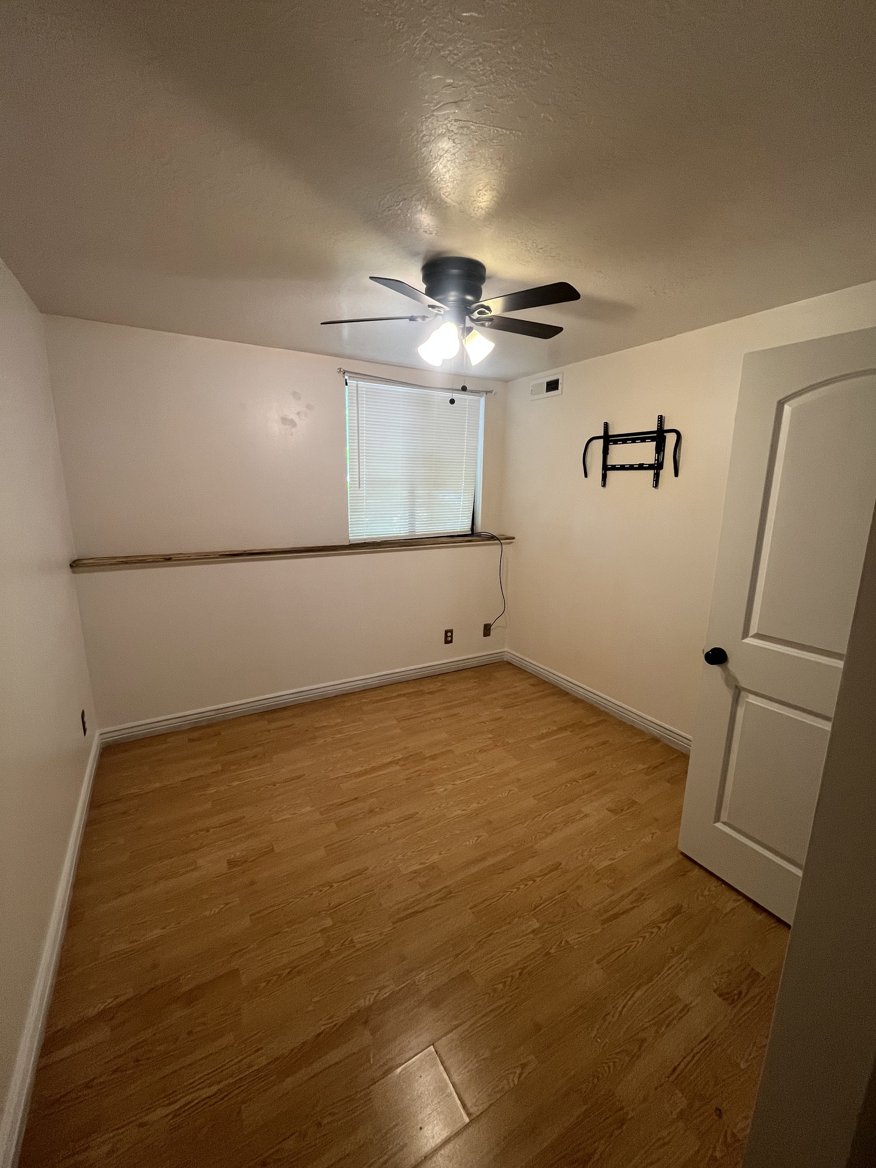 Room featuring wood-finish flooring, a ceiling fan with integrated lighting, a window with horizontal blinds, and a paneled door with black hardware