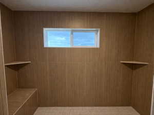 Primary ensuite walk-in shower featuring patterned tile flooring, wood-look wall tile, dual shower heads, a built-in shower bench, a corner shelf, and a frosted transom window