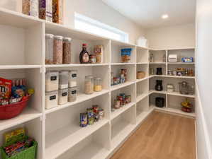 Spacious walk-in pantry featuring extensive white shelving, wood-finish flooring, recessed lighting, and a high window