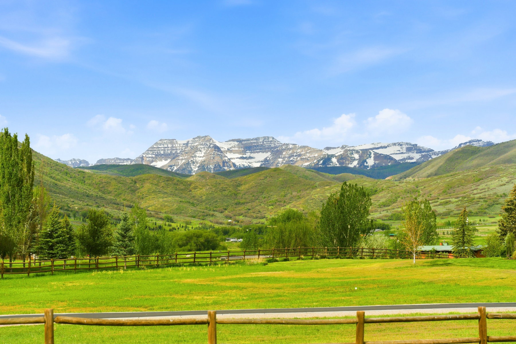 Expansive green field bordered by a rustic wooden fence, featuring distant snow-capped mountains and rolling green hills