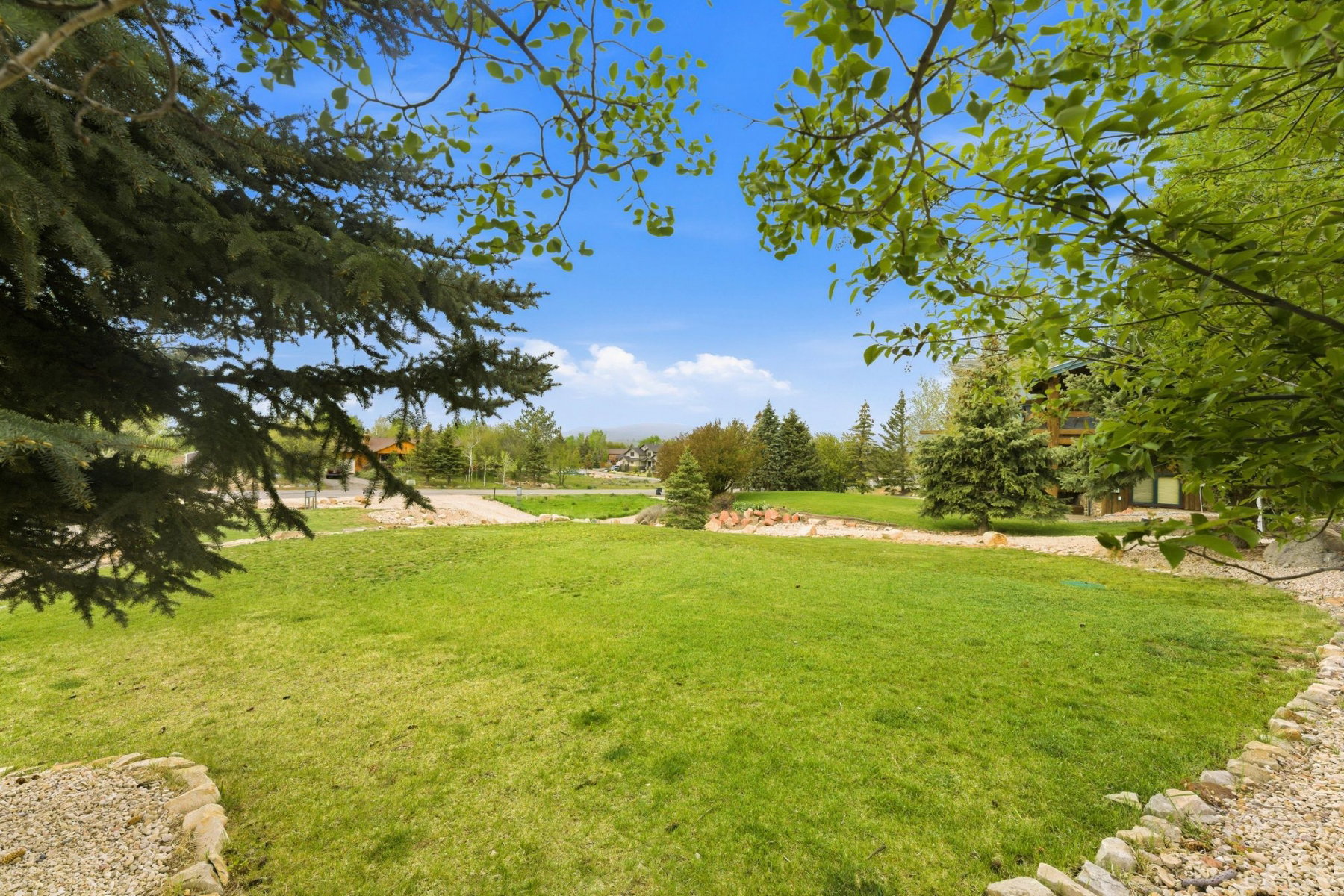 Expansive lawn area surrounded by mature trees, featuring a stone-edged perimeter and distant residential structures