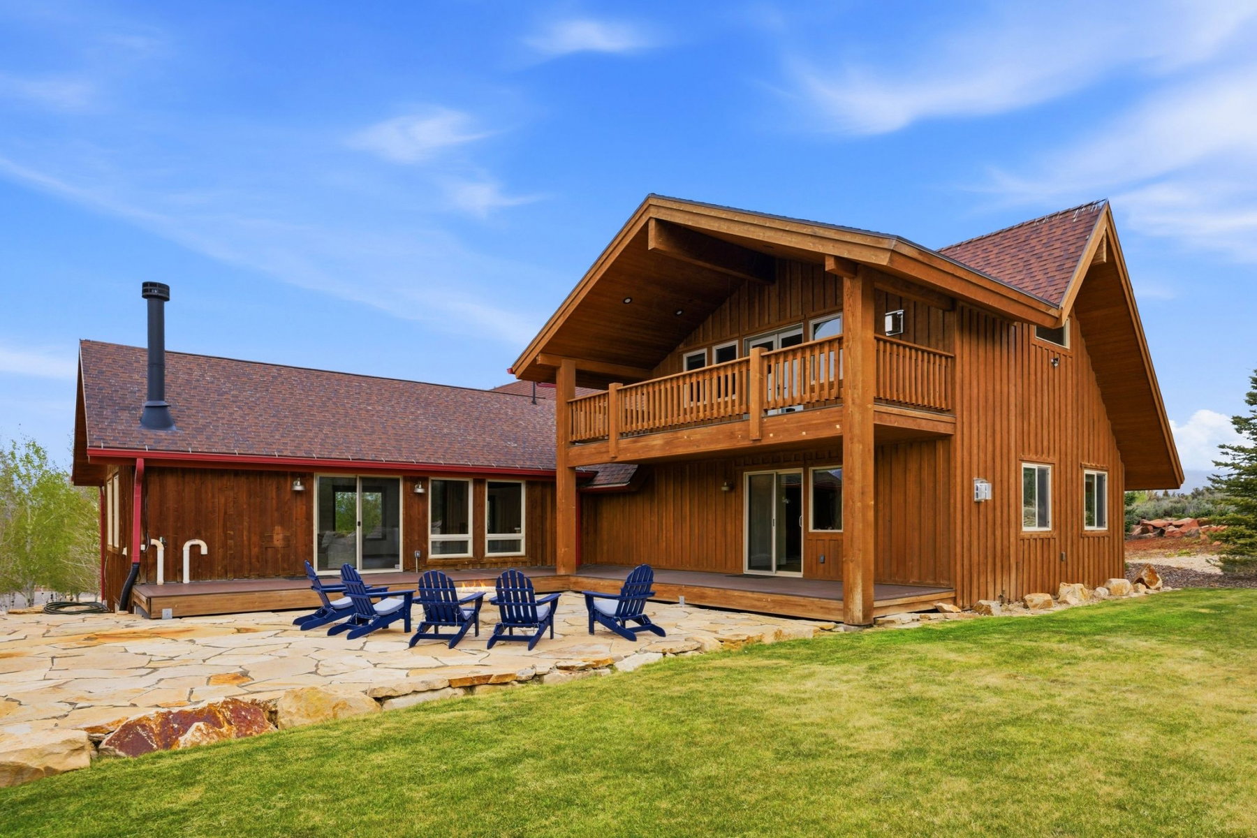 Wood-sided residence featuring a large stone patio, a second-story balcony, multiple sliding glass doors, and a verdant lawn