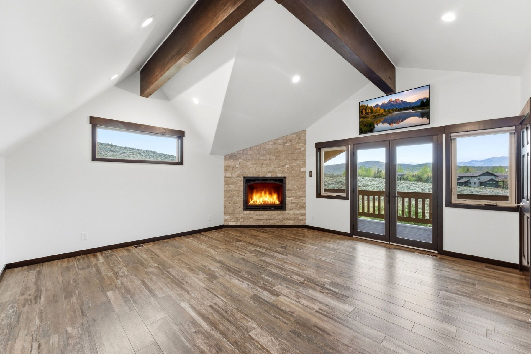 Spacious room featuring vaulted ceilings with exposed wood beams, wood-finish flooring, a stone-clad fireplace, recessed lighting, and multiple windows with dark trim