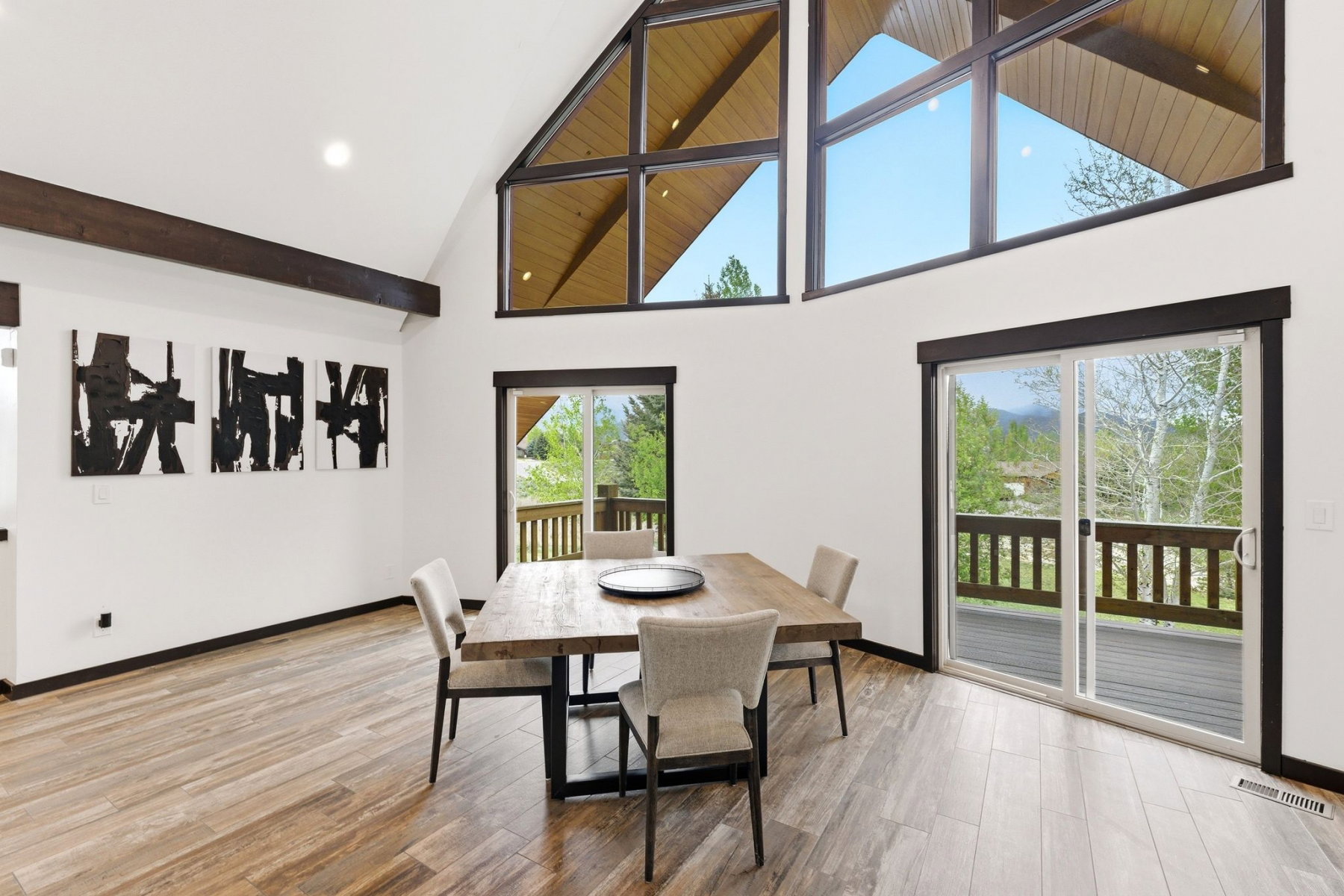 Vaulted ceiling with exposed beams, expansive window array, wood-finish flooring, and sliding glass doors opening to a wood deck