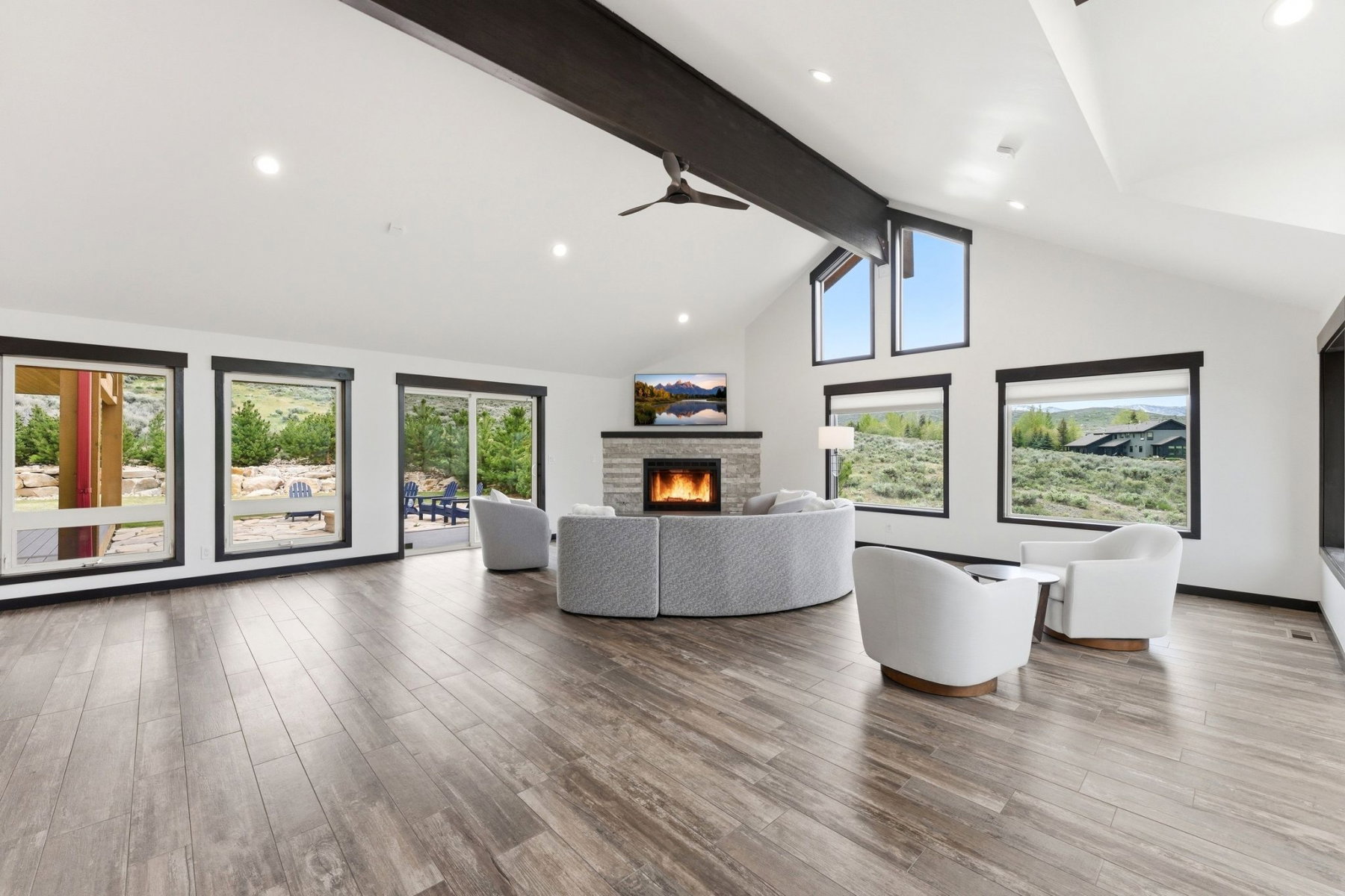 Expansive living space featuring wood-finish flooring, a stone fireplace with a hearth, vaulted ceilings with exposed dark beams, and multiple large windows with dark trim