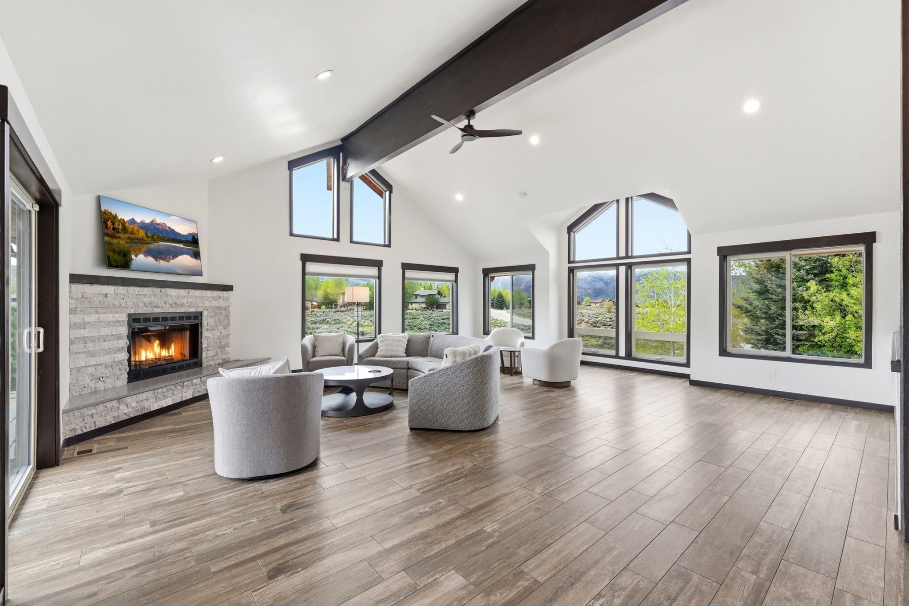 Spacious living area featuring a vaulted ceiling with exposed beam, a stone fireplace, wood-finish flooring, and multiple large windows
