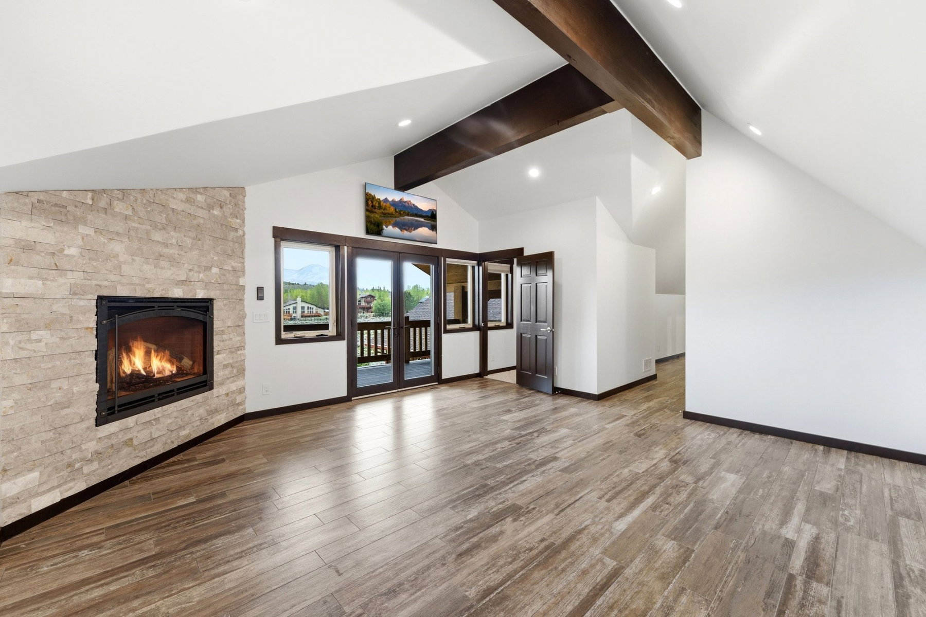 Spacious room featuring vaulted ceilings with exposed wood beams, a stone-clad fireplace, wood-finish flooring, recessed lighting, and balcony access through double glass doors