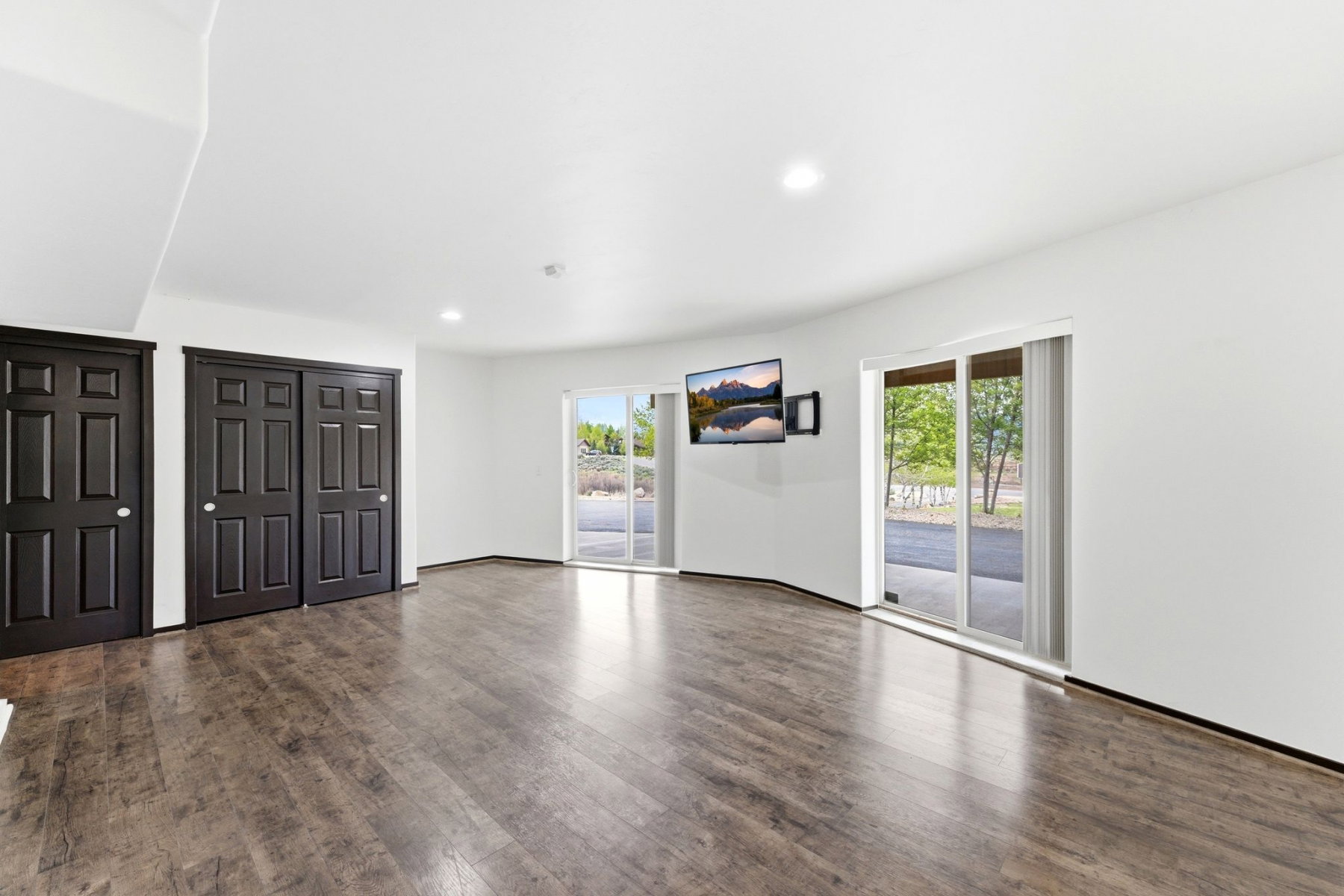 Spacious room featuring wood-finish flooring, two sets of dark paneled doors, and two sets of sliding glass doors