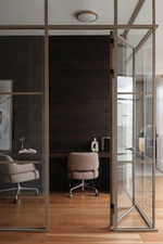 Glass partition walls with a hinged entry, integrated dark wood-finish desk, wood-finish flooring, and a flush-mount ceiling light