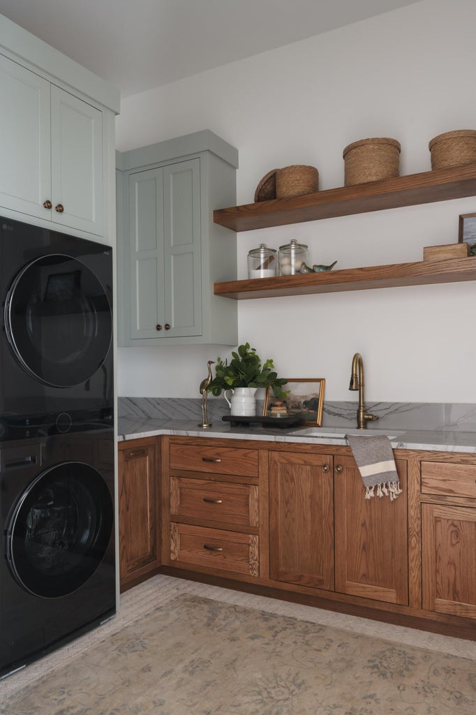 Dedicated laundry area featuring stacked appliances, custom wood-finish cabinetry, a single basin sink with a brass-finish faucet, floating wood shelves, and a stone countertop with a matching backsplash