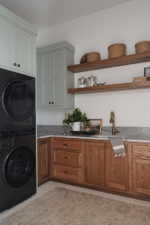 Dedicated laundry area featuring stacked appliances, custom wood-finish cabinetry, a single basin sink with a brass-finish faucet, floating wood shelves, and a stone countertop with a matching backsplash