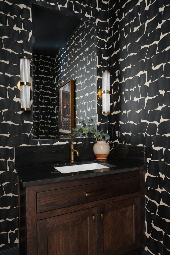 Powder room with patterned wallpaper, a dark wood vanity, black countertop, rectangular undermount sink, and brass-finish faucet