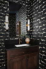 Powder room with patterned wallpaper, a dark wood vanity, black countertop, rectangular undermount sink, and brass-finish faucet