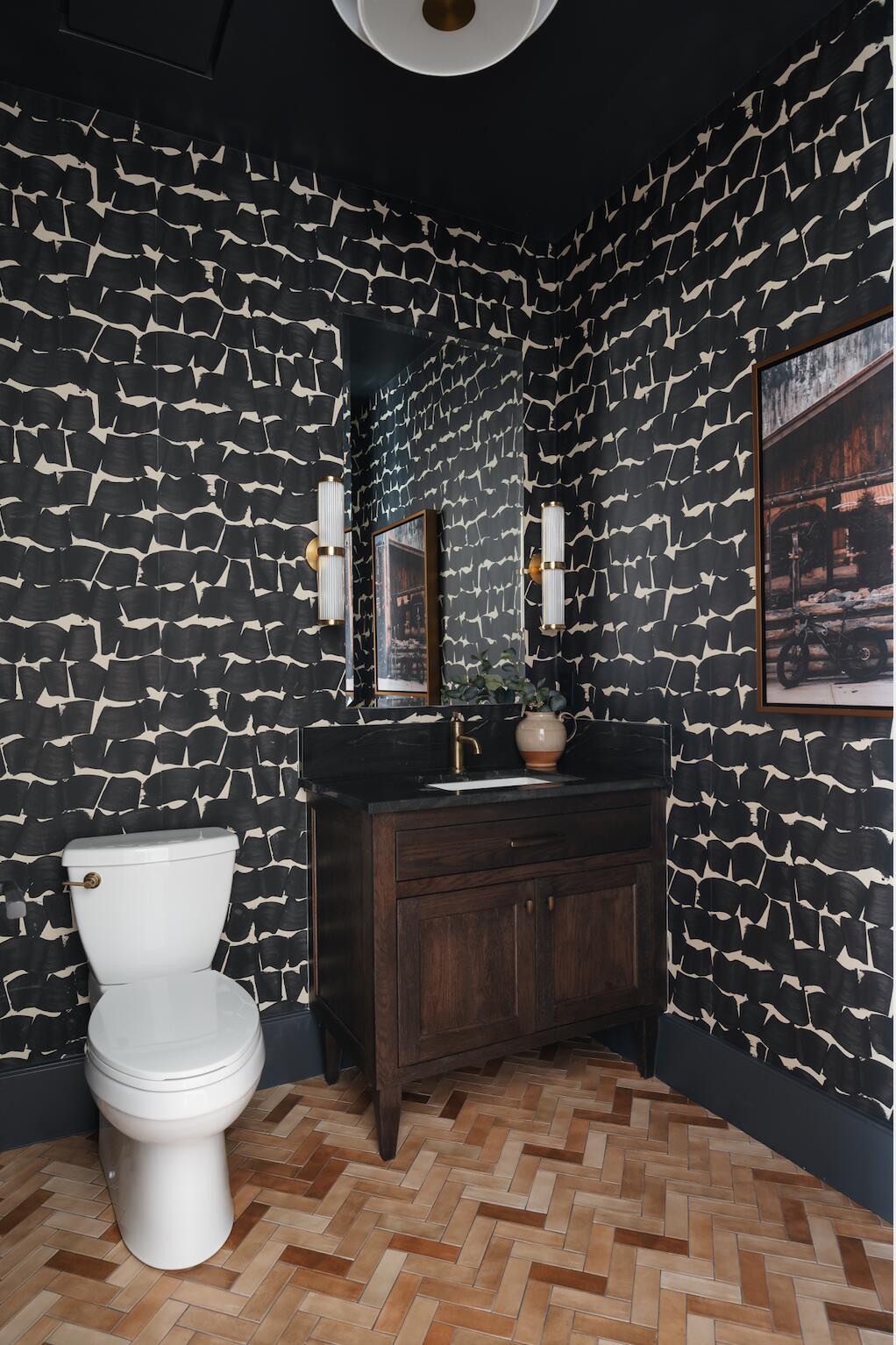 Powder room featuring striking patterned wall covering, a dark wood-finish vanity with a dark countertop, brass-finish fixtures, and herringbone wood-finish flooring