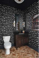 Powder room featuring striking patterned wall covering, a dark wood-finish vanity with a dark countertop, brass-finish fixtures, and herringbone wood-finish flooring