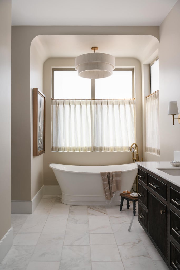 Freestanding soaking tub set within a windowed alcove, featuring a tiered drum chandelier, white tiled flooring, and a dark wood vanity with a white countertop