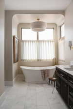 Freestanding soaking tub set within a windowed alcove, featuring a tiered drum chandelier, white tiled flooring, and a dark wood vanity with a white countertop
