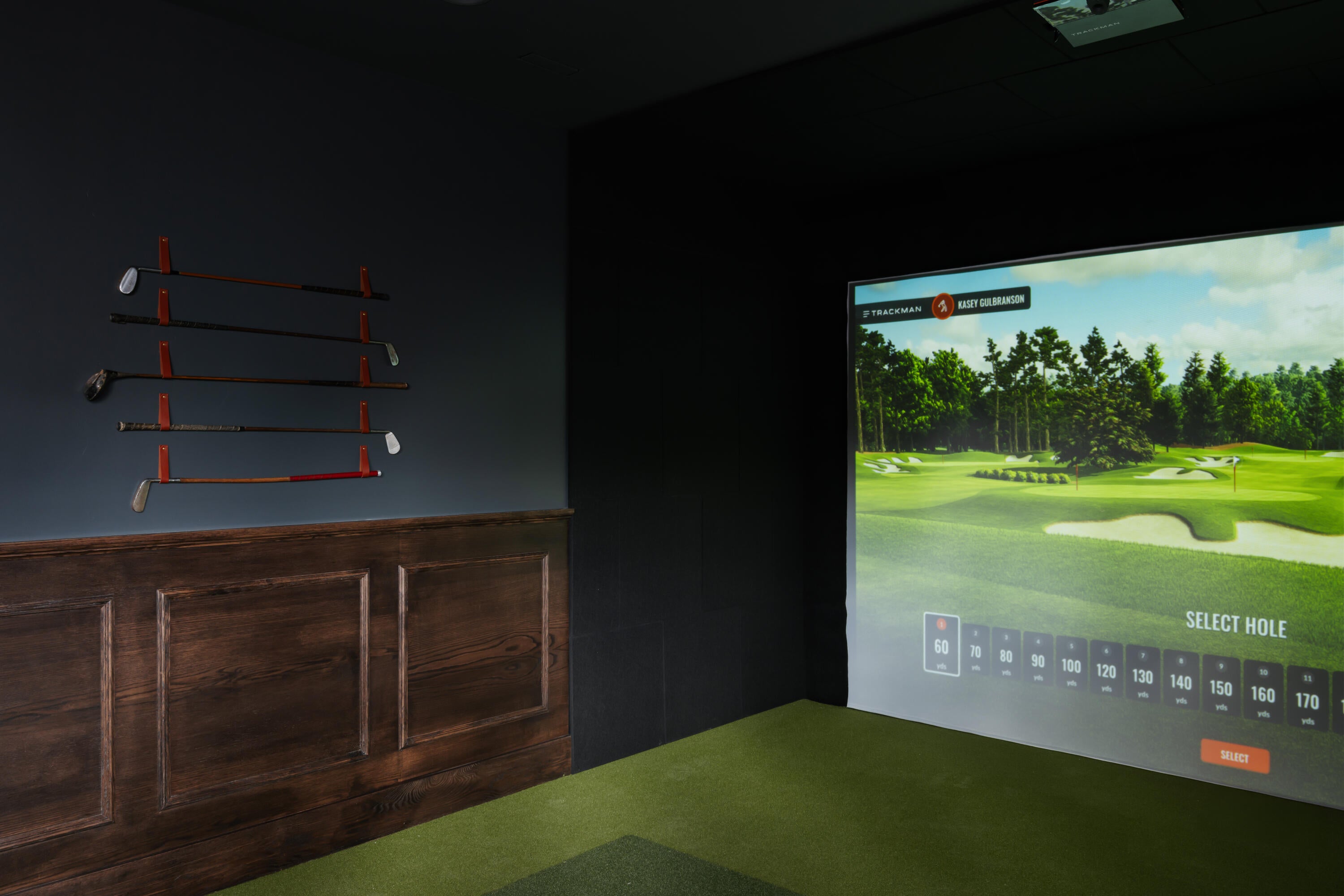 Dedicated golf simulator room featuring a large projection screen, dark wall finishes, and a green turf floor