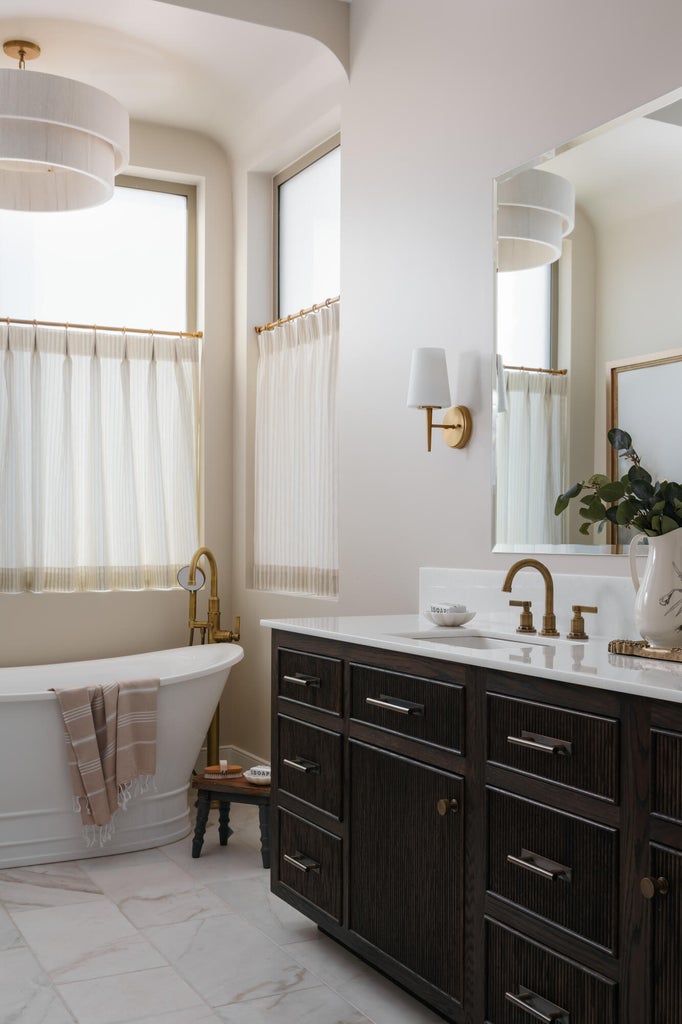 Expansive bathroom featuring a freestanding soaking tub with floor-mounted brass fixtures, a dark wood-finish vanity with ample storage, a white countertop, and brass wall sconces