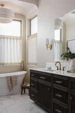 Expansive bathroom featuring a freestanding soaking tub with floor-mounted brass fixtures, a dark wood-finish vanity with ample storage, a white countertop, and brass wall sconces
