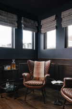 Dark-toned interior space featuring wood-finish flooring, dark wall paneling, and multiple windows with Roman shades