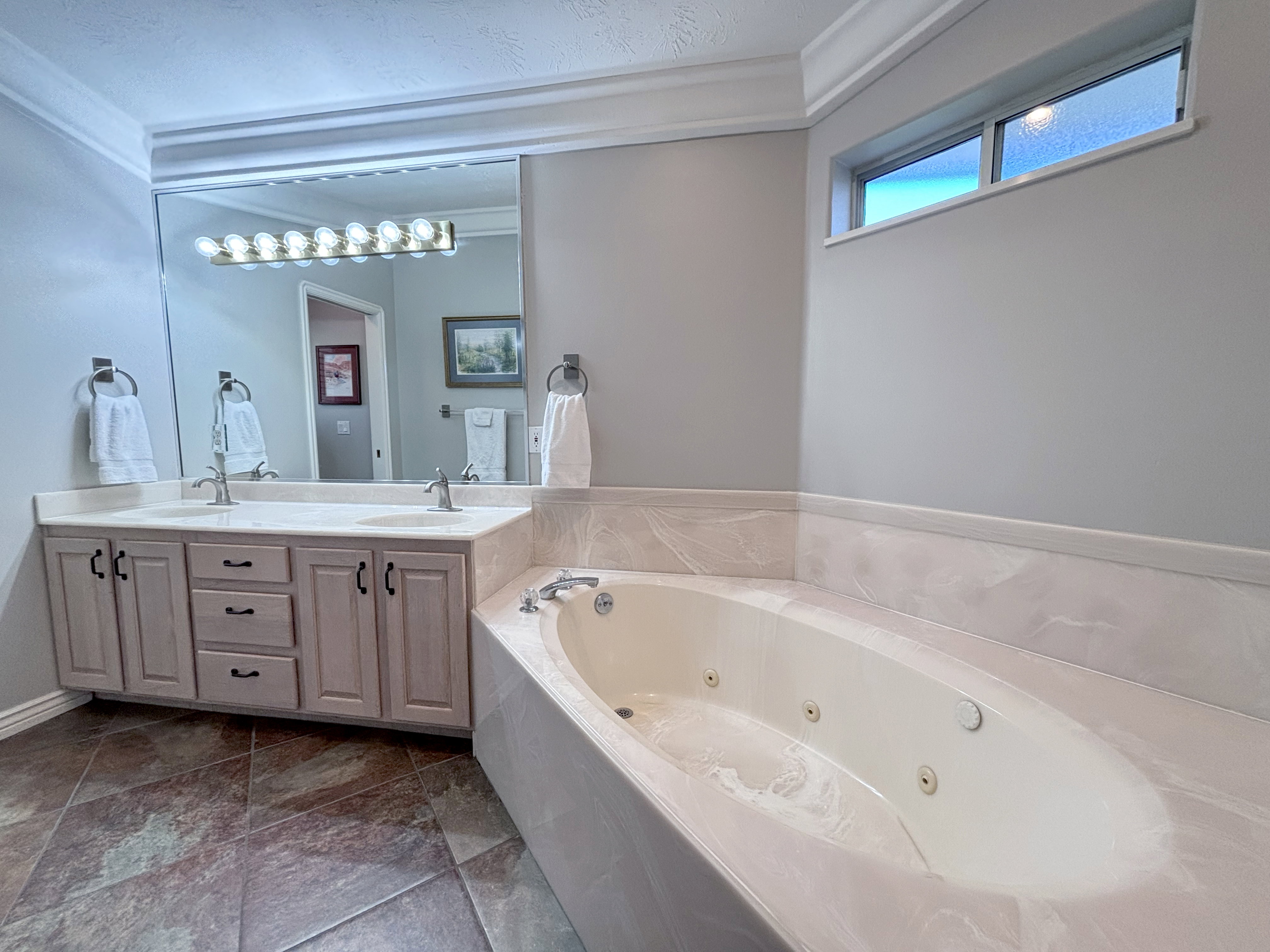 Jetted soaking tub with surrounding stone-finish decking, dual-sink vanity with stone-finish countertop, large frameless mirror, and transom window