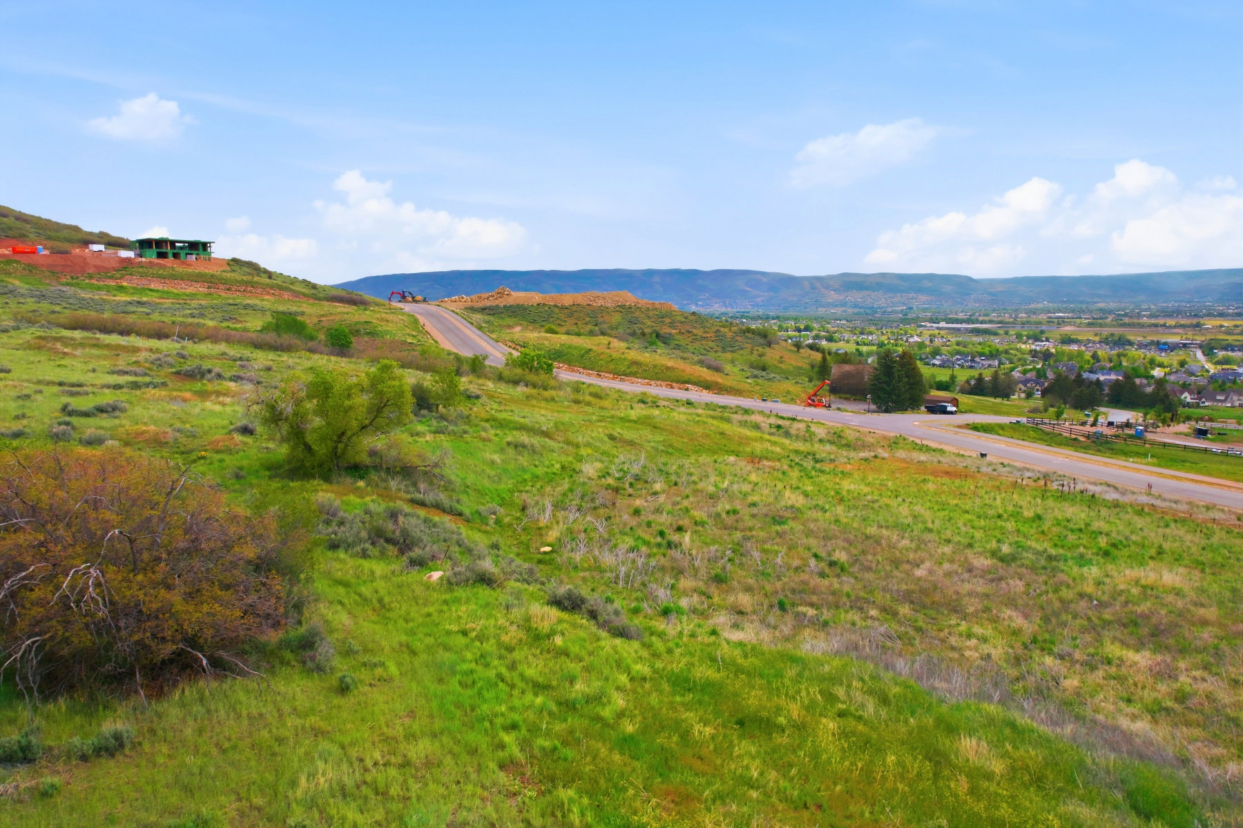 Expansive sloping terrain featuring a paved road, scattered green shrubs, and distant residential developments