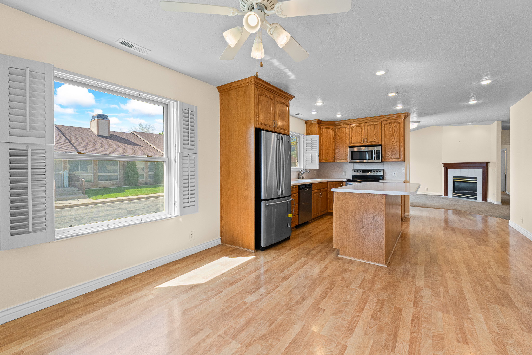 Open-concept living space with wood-finish flooring, a kitchen featuring wood cabinetry and stainless steel appliances, a center island with a white countertop, recessed lighting, and a living area with a fireplace