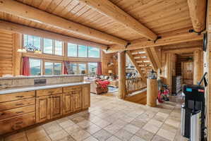Great room featuring expansive window walls, natural wood log construction, exposed beam ceilings, a built-in kitchen peninsula with a light-toned countertop, and tiled flooring