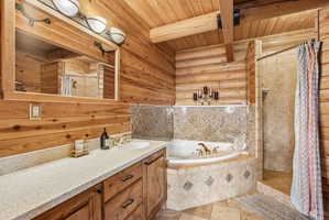 Bathroom featuring wood-paneled walls, a double vanity with an integrated sink, a jetted corner tub with tiled surround, a tiled shower, and exposed wood ceiling beams