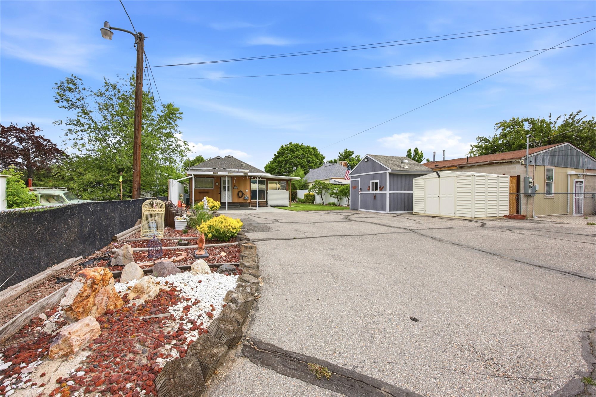 Paved driveway with dedicated parking, featuring a detached garage with gray siding and white trim, an additional metal storage shed, and a separate building with a flat roof