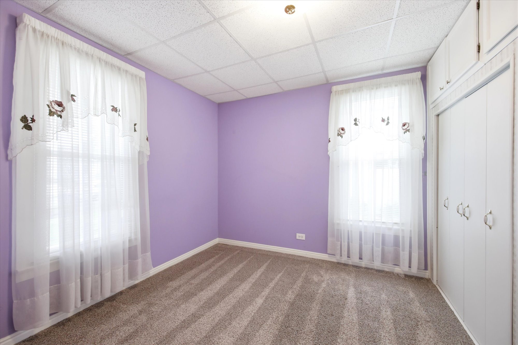 Carpeted room featuring two windows with blinds and sheer curtains, overhead lighting, and built-in white cabinetry with closed storage