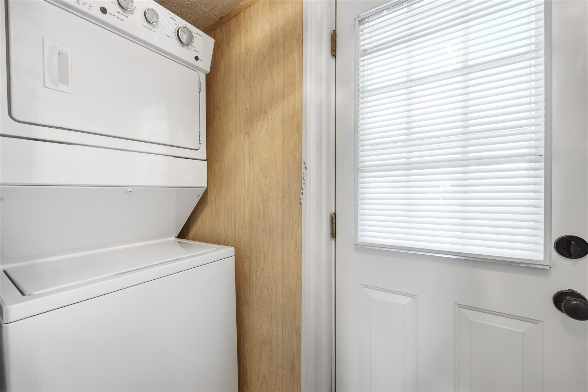 Stackable washer and dryer unit with a light wood-finish paneled wall and a white door featuring an inset window with blinds