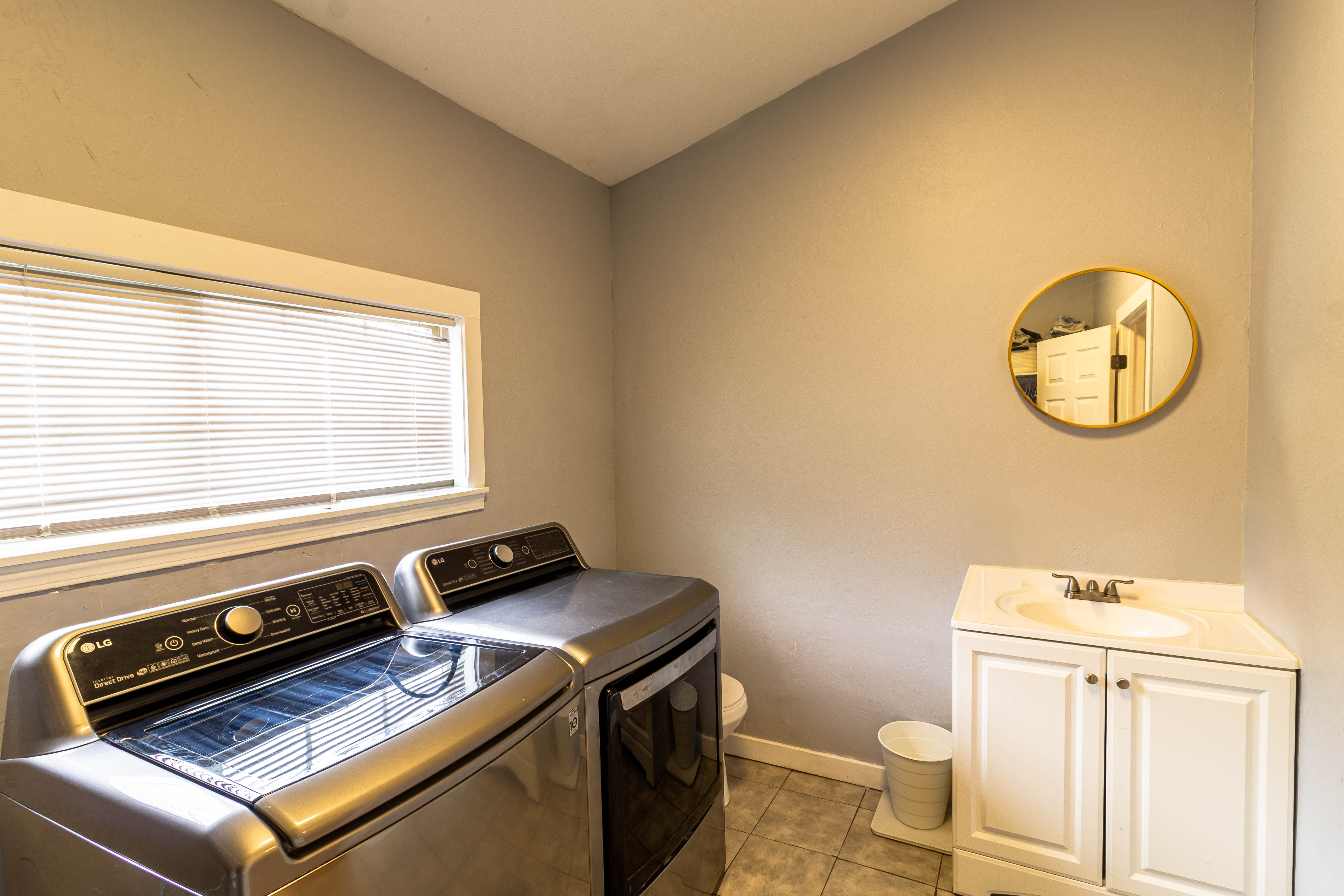 Utility room featuring a window with blinds, a white vanity with a basin sink, an oval mirror with a gold frame, and square floor tiling