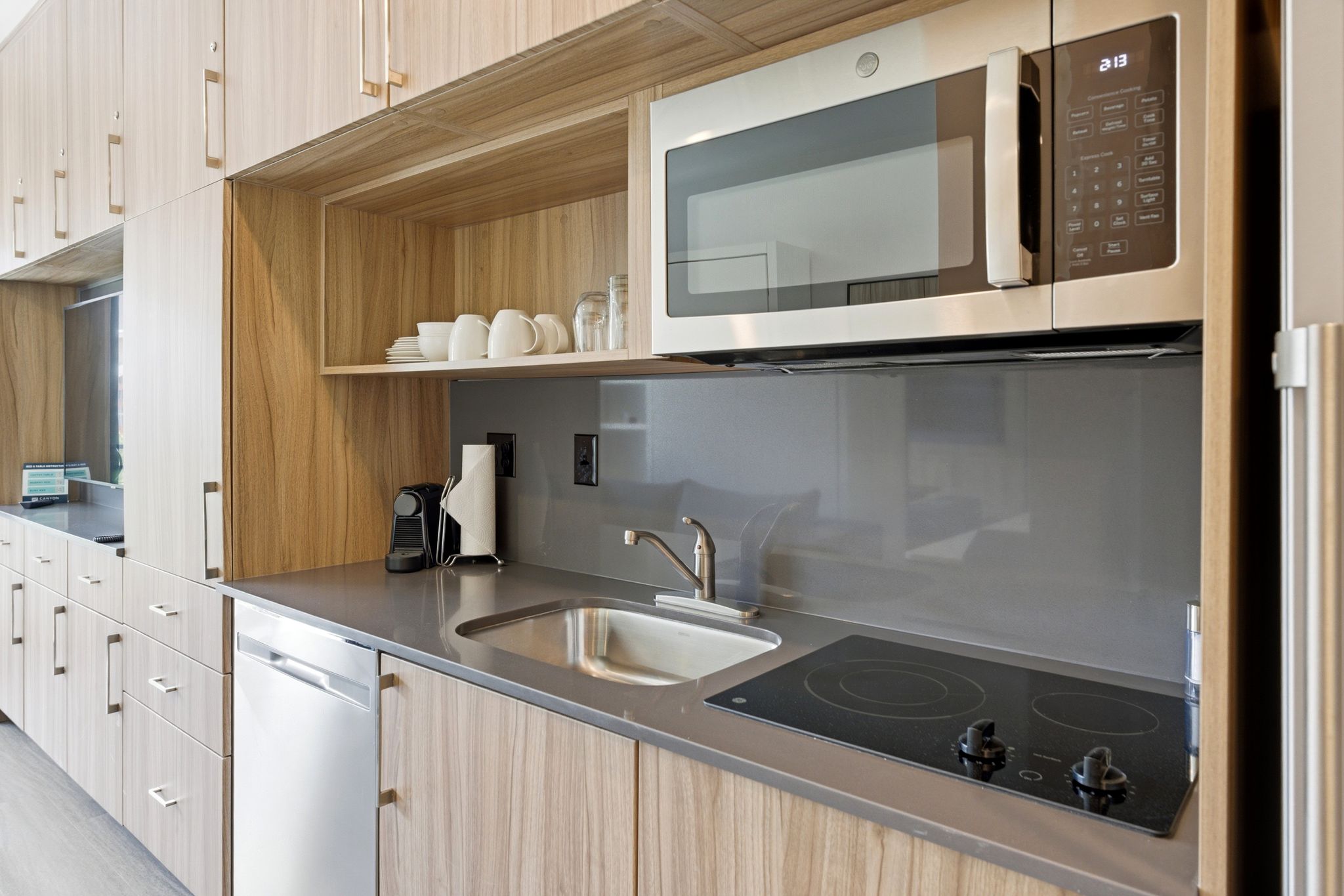 Compact kitchen featuring wood-grain cabinetry, stainless steel microwave, integrated cooktop, undermount sink, and a stainless steel dishwasher