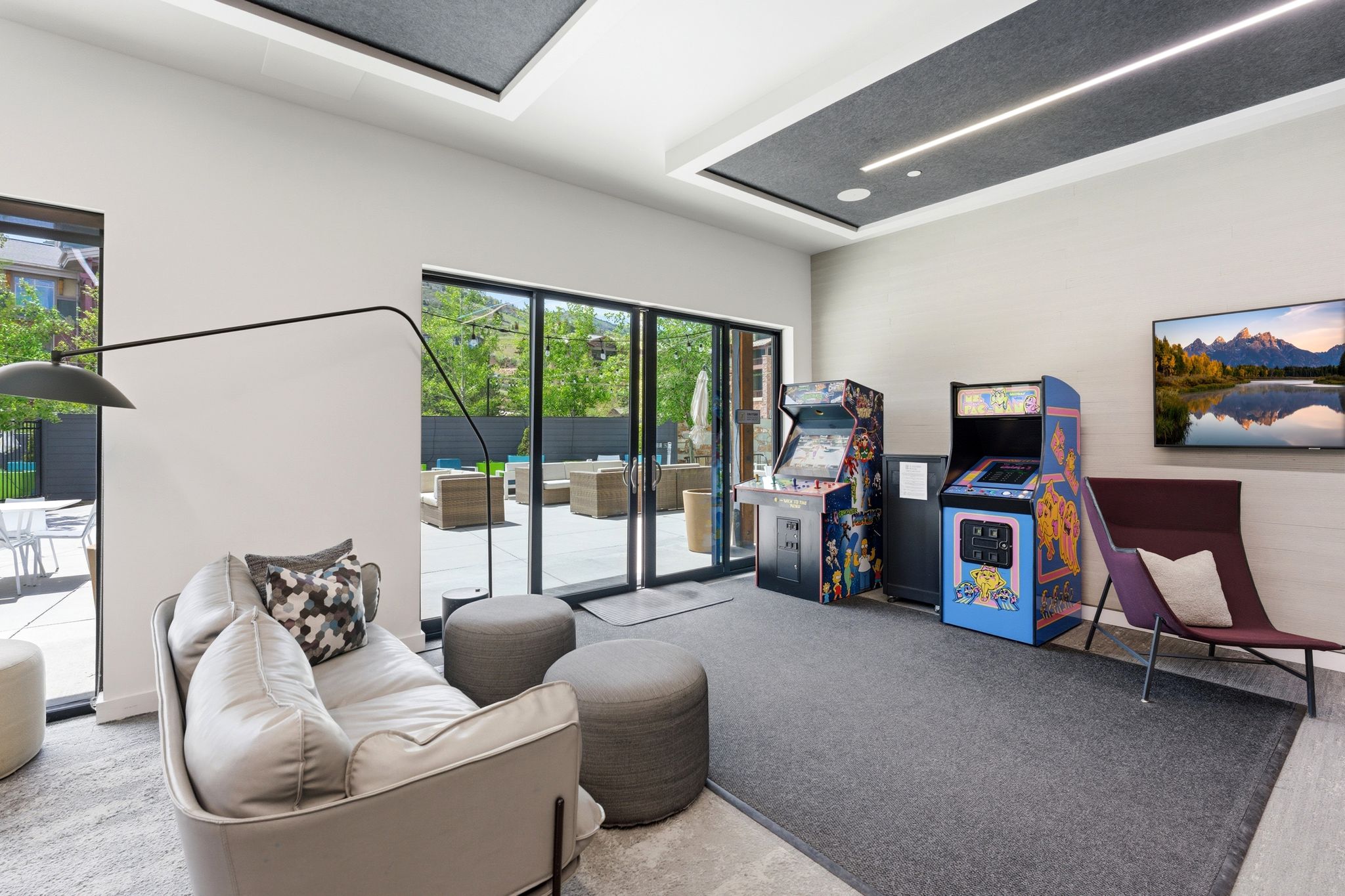 Spacious recreation area featuring a wall-mounted display, recessed ceiling lighting, and large glass doors opening to an outdoor patio
