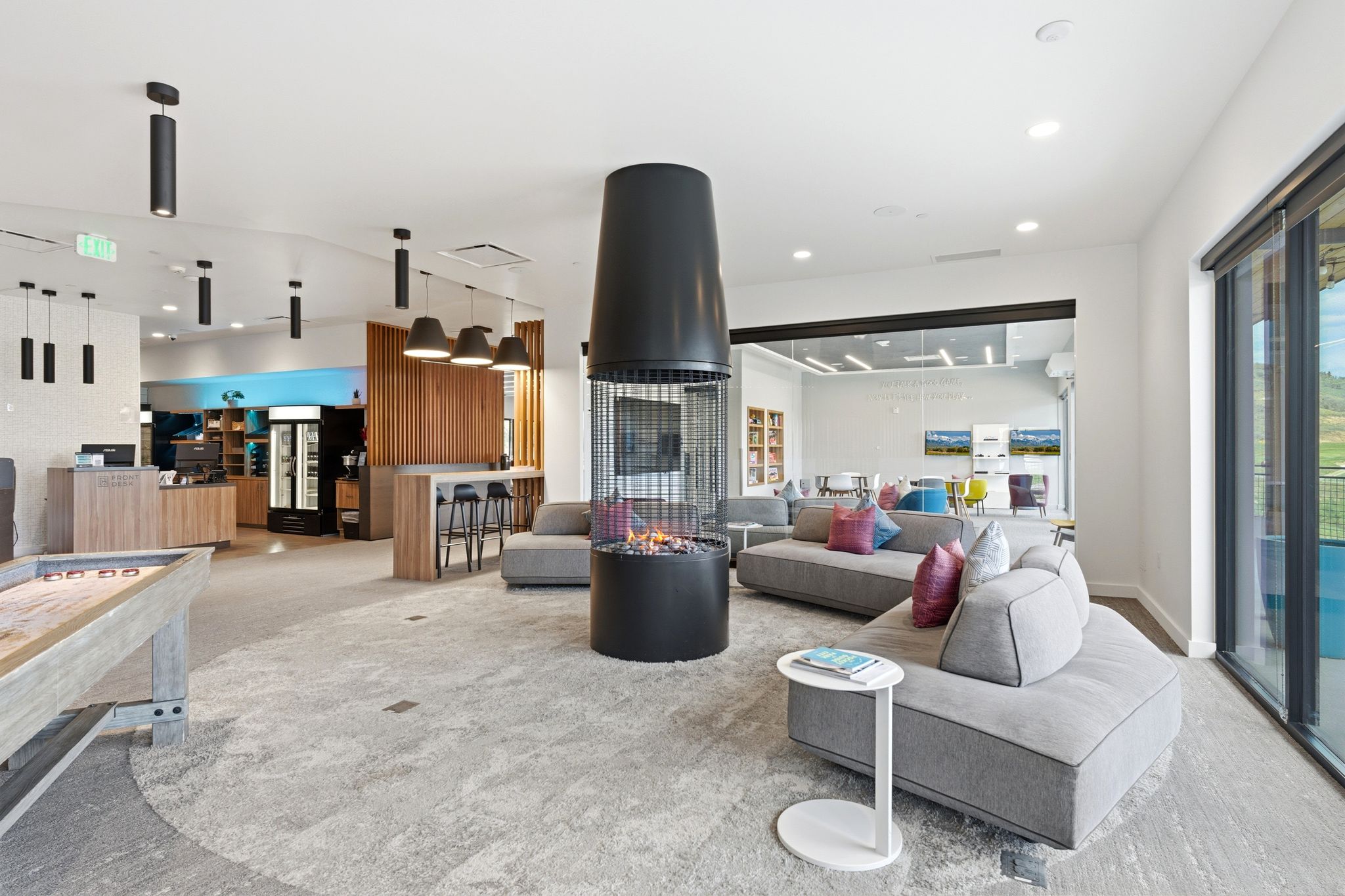 Spacious common area featuring a central cylindrical fireplace, a modern bar area with built-in cabinetry, large format glass doors, and recessed lighting throughout