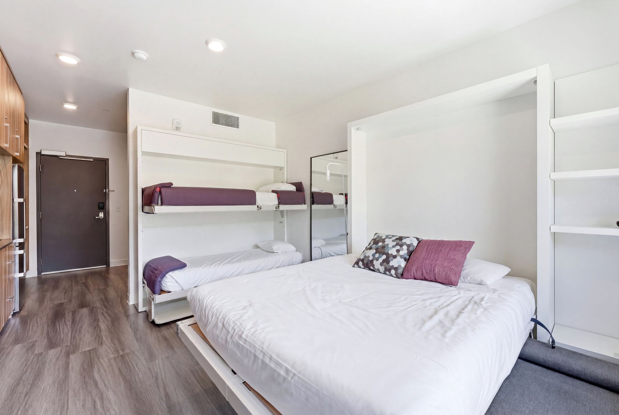 Studio living area featuring a wall-mounted bunk bed system, additional wall-mounted bed, built-in shelving, wood-finish flooring, and recessed lighting
