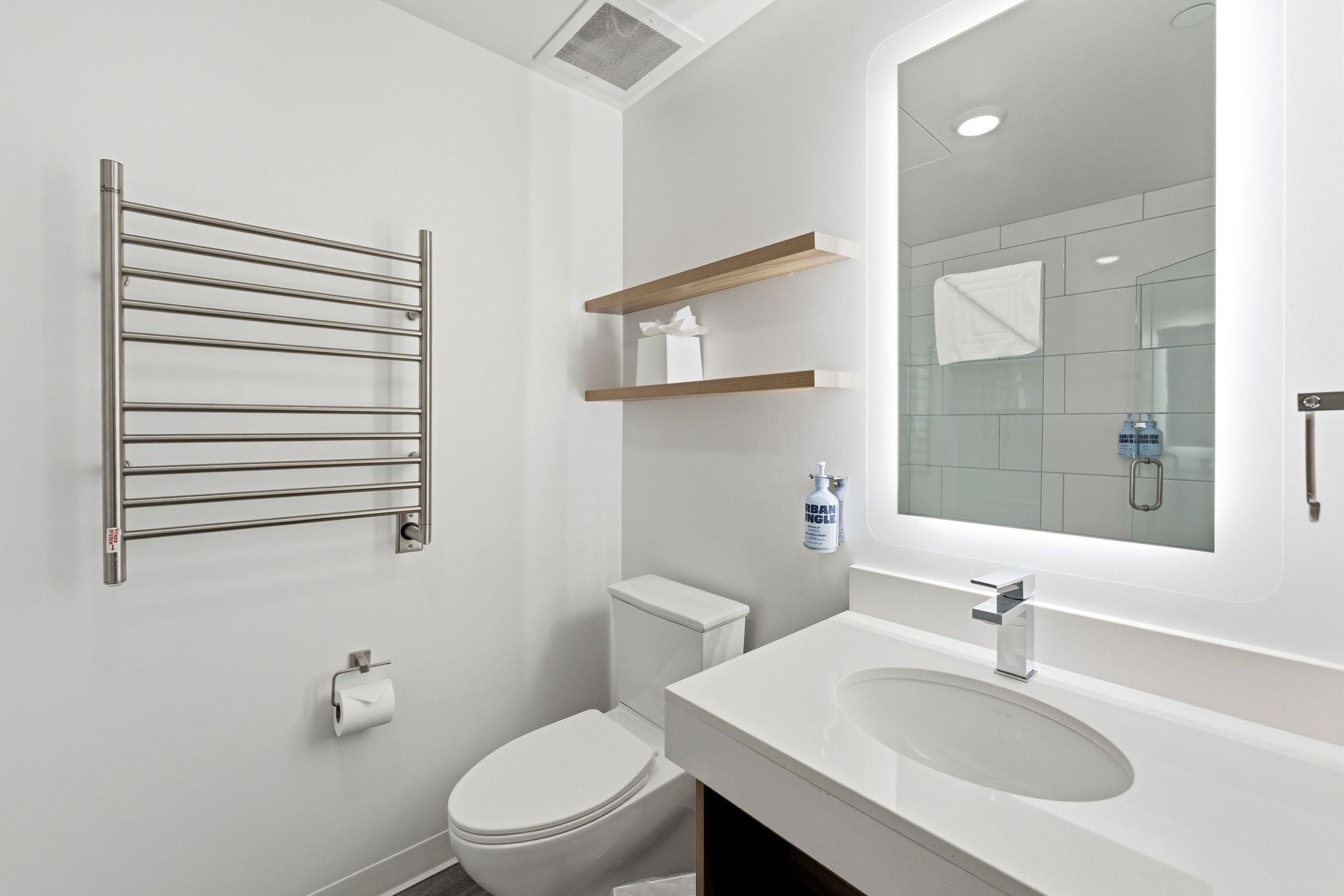 Modern bathroom featuring a vanity with an integrated sink, illuminated mirror, wall-mounted towel warmer, and floating wood-finish shelving